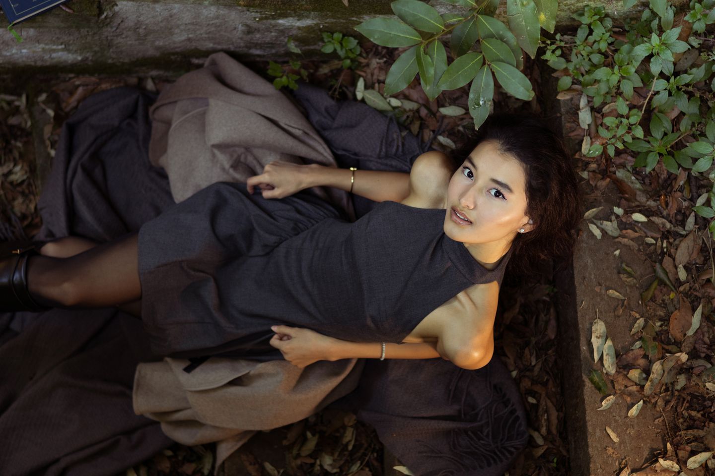 A woman lies among fallen leaves and moss-covered stone steps in a dark sleeveless dress, a brown wool coat draped beside her, reaching one arm up and looking directly at the camera.
