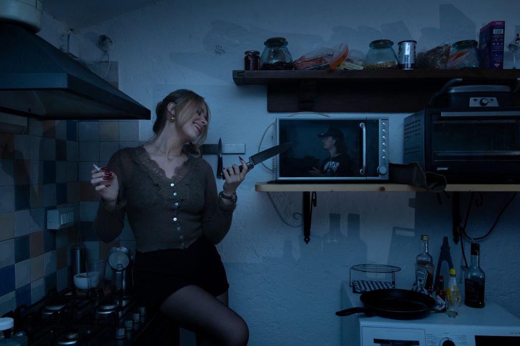 A woman leans against a kitchen counter at night under cold blue light, laughing while holding a lit cigarette and a kitchen knife, a photo of a man visible on a microwave screen behind her.