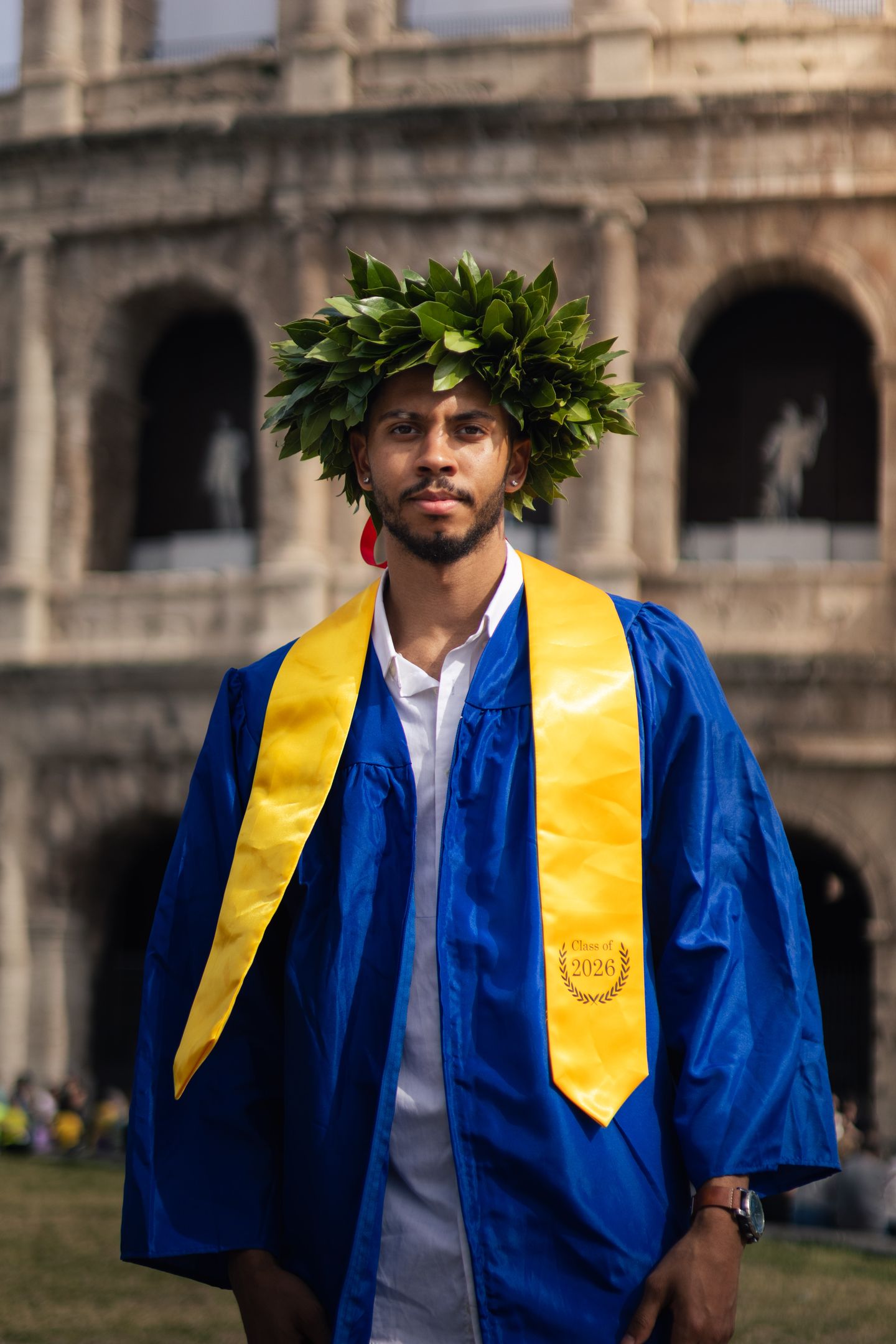 A young man in a blue graduation gown and gold sash, wearing a laurel wreath, stands before the arched facade of the Colosseum.
