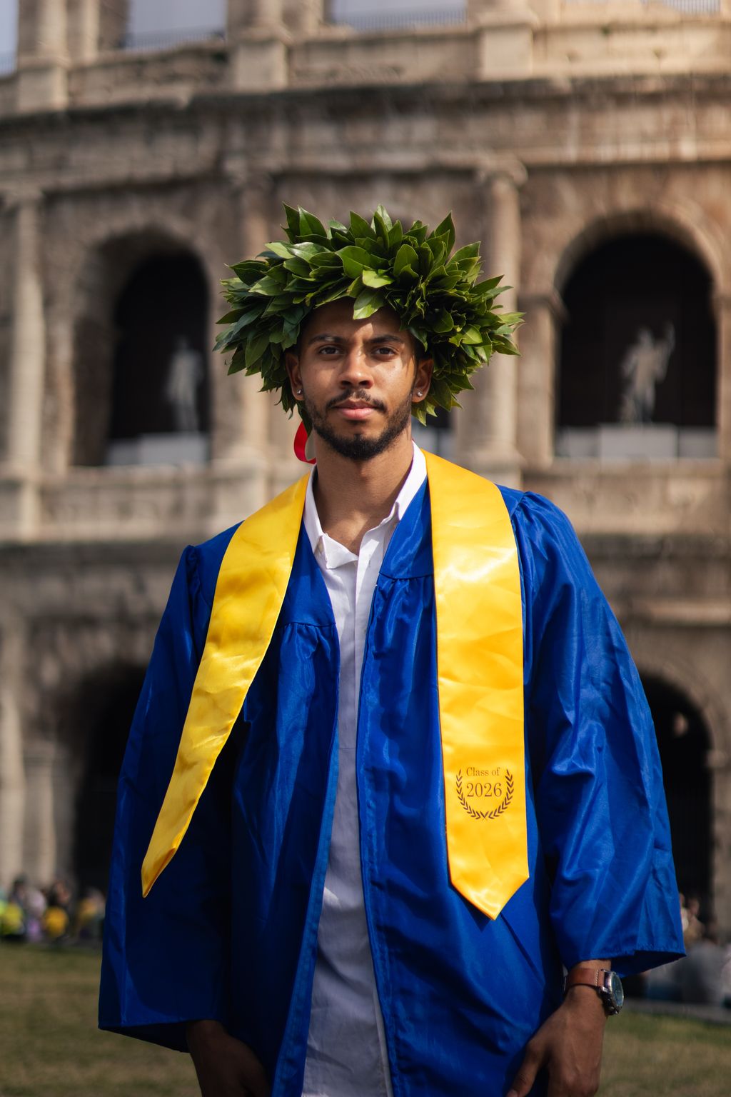 A young man in a blue graduation gown and gold sash, wearing a laurel wreath, stands before the arched facade of the Colosseum.