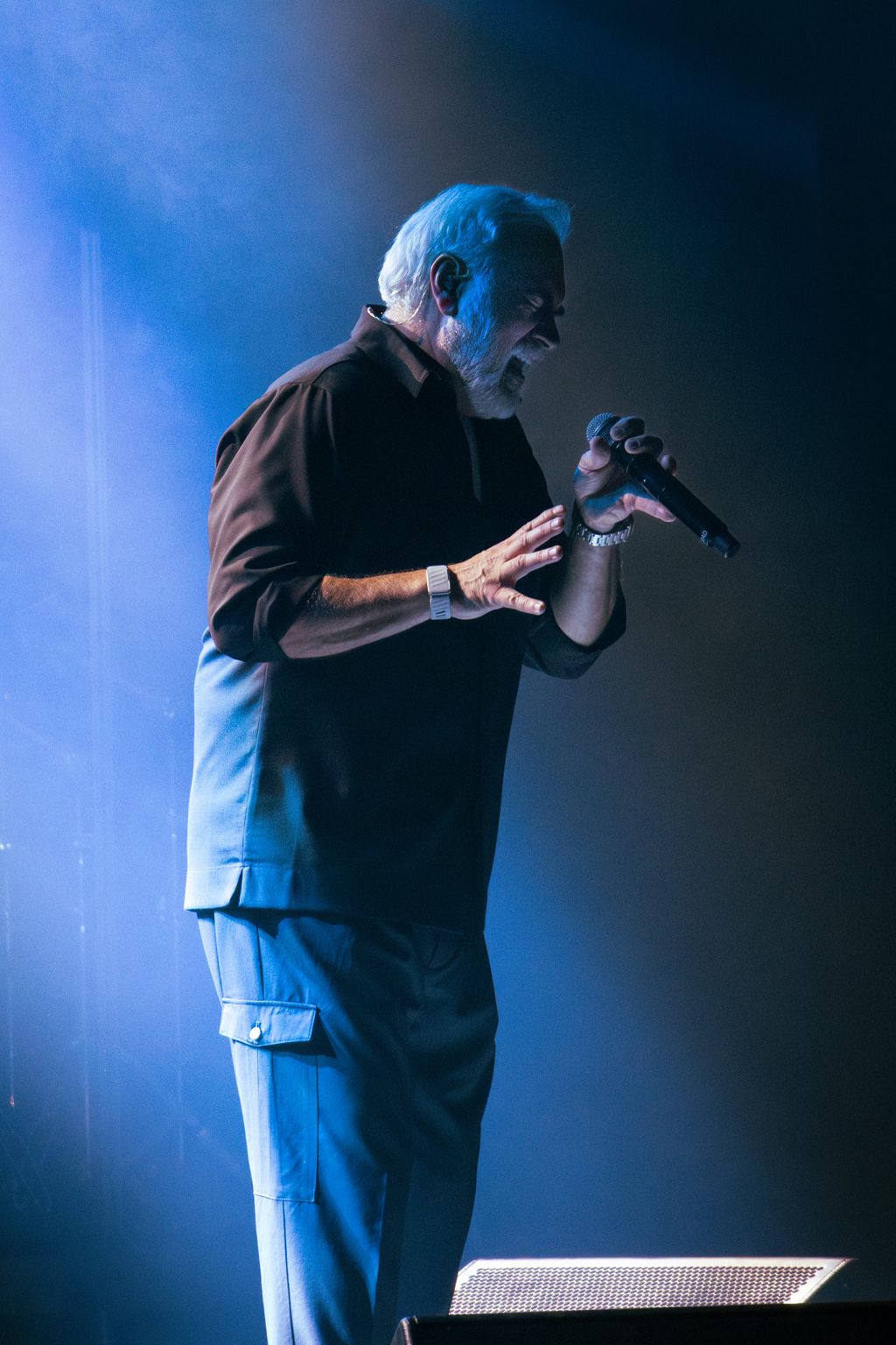 Side profile of an older man singing into a microphone on a smoky, blue-lit concert stage, one hand raised.