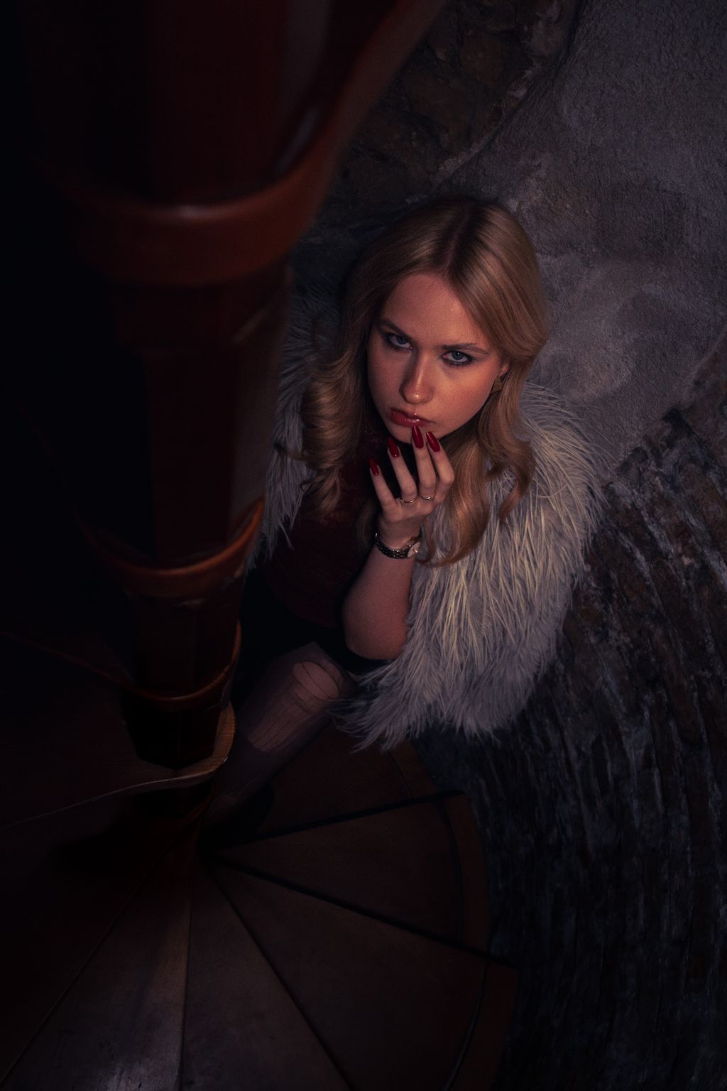 Overhead portrait of a blonde woman in a faux-fur wrap, crouched partway down a dim wooden spiral staircase, resting her chin on her hand.