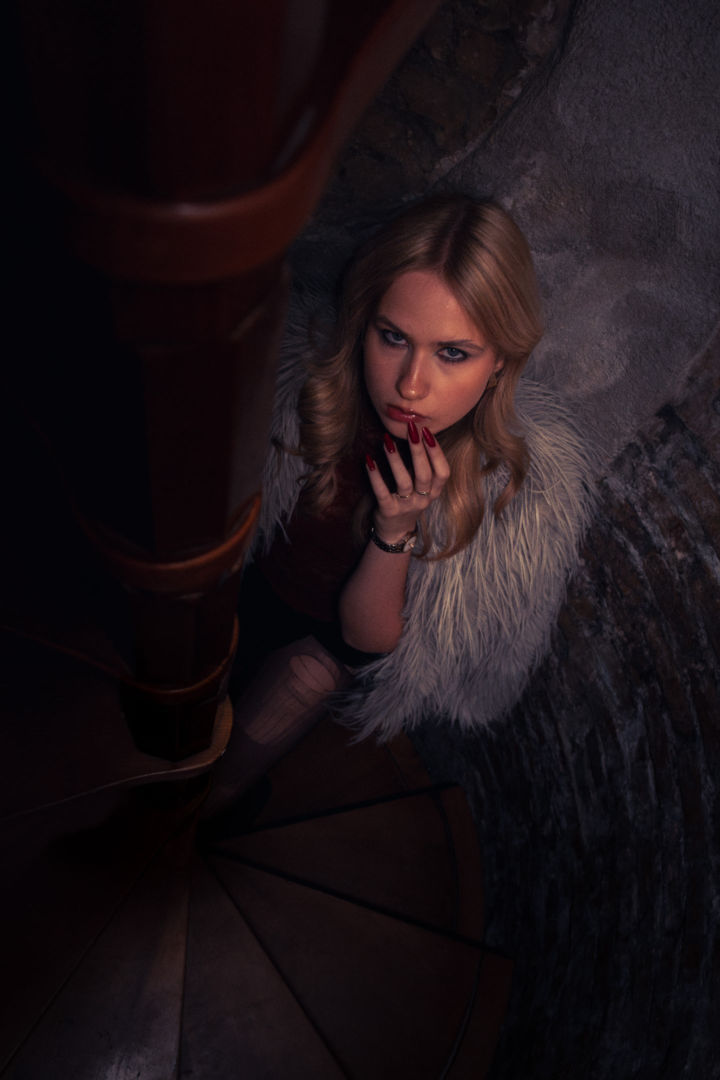 Overhead portrait of a blonde woman in a faux-fur wrap, crouched partway down a dim wooden spiral staircase, resting her chin on her hand.