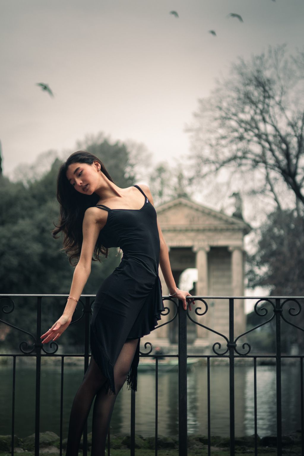 A young woman in a black tie and white collared shirt leans against a stone parapet above a road at dusk, looking back over her shoulder as car headlights pass below.