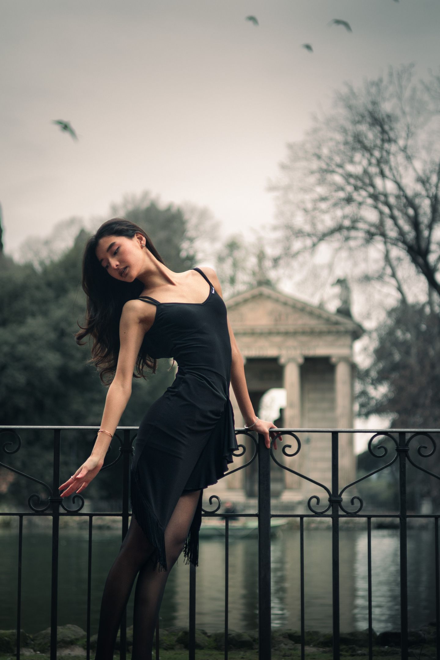 A young woman in a black tie and white collared shirt leans against a stone parapet above a road at dusk, looking back over her shoulder as car headlights pass below.