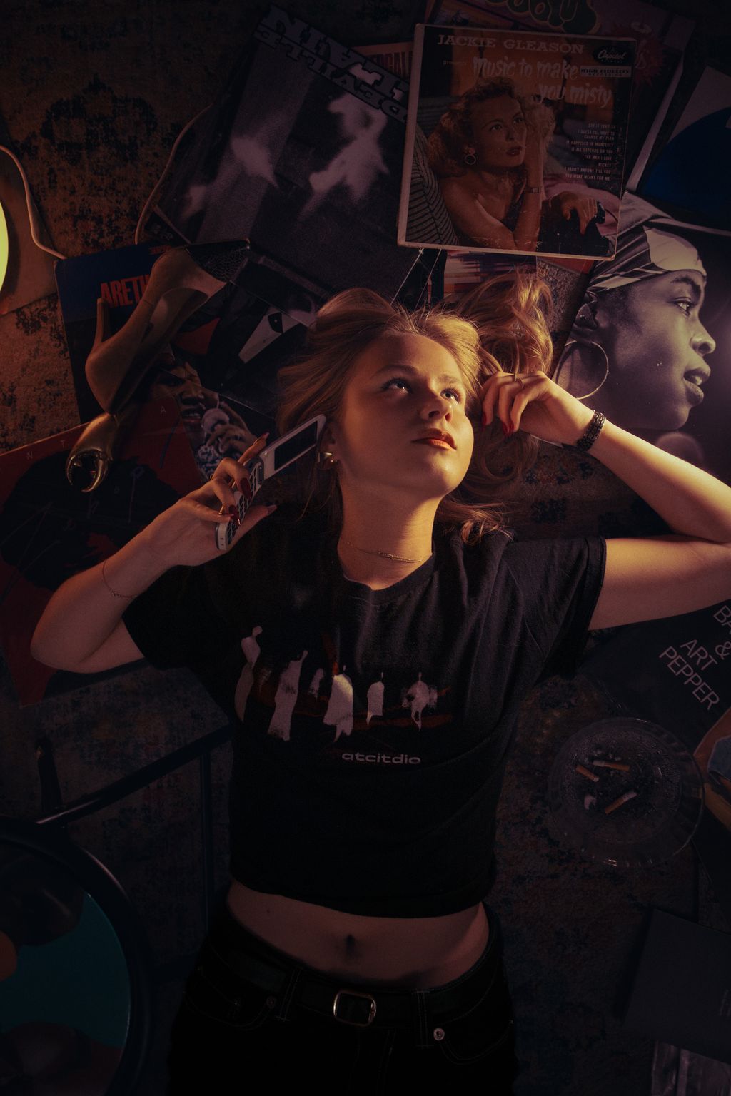 Overhead shot of a young woman lying atop a stack of vintage vinyl records, talking on a corded phone, surrounded by album covers and an ashtray.